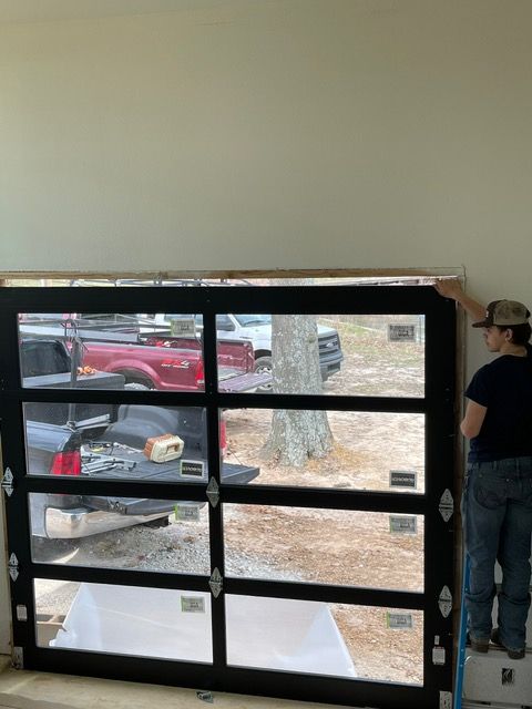 Person installing a black-framed glass garage door. Daylight shines through, showing vehicles outside.