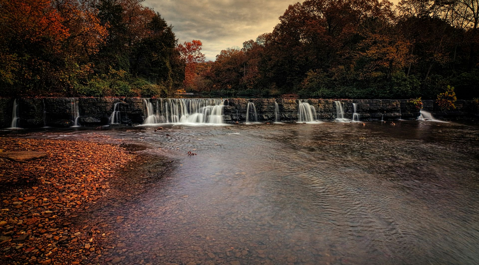 Autumn waterfall over a calm river, with orange leaves and trees along the banks.