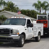 White utility truck with small U.S. flag parked in front of a house, carrying an orange skid steer loader.