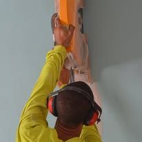 Worker using a trowel to apply orange material to a wall while wearing red ear защитors and a yellow shirt