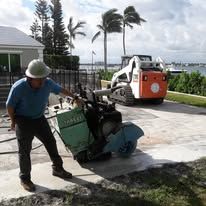 Worker using a concrete saw on a driveway near the waterfront, with a skid steer in the background.
