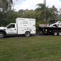 White service truck towing a black trailer with equipment in a grassy yard, trees in the background