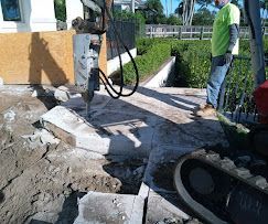 Damaged sidewalk and broken concrete beside a roadside work site, with a worker standing nearby