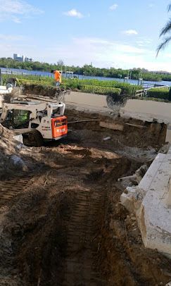A Bobcat excavator digs a trench near a waterfront; a worker in orange vest oversees.