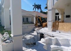 Construction debris and an excavator at a damaged white building with palm trees nearby