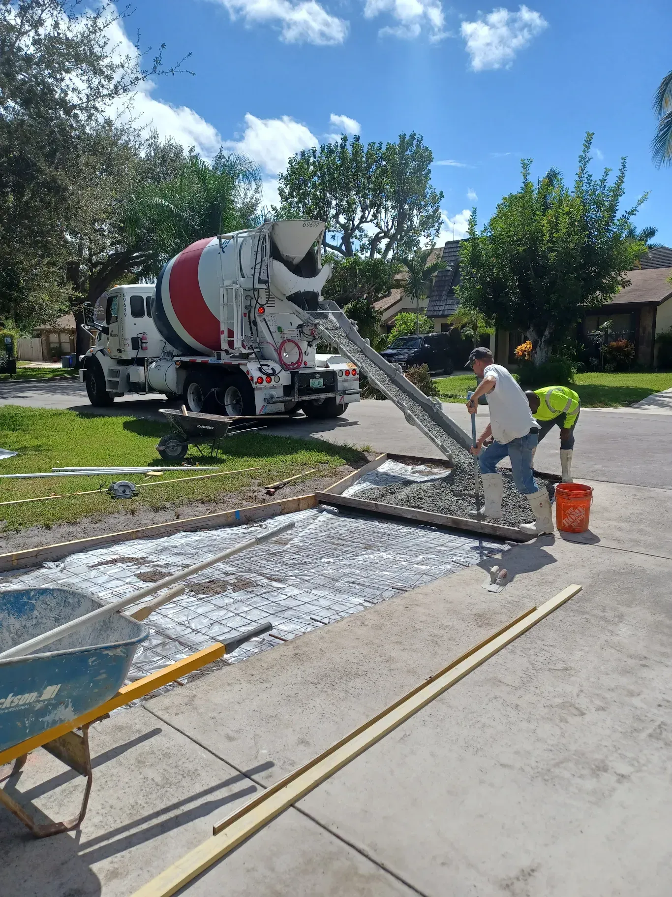 Concrete truck pouring a driveway slab while a worker smooths wet concrete in a sunny neighborhood.