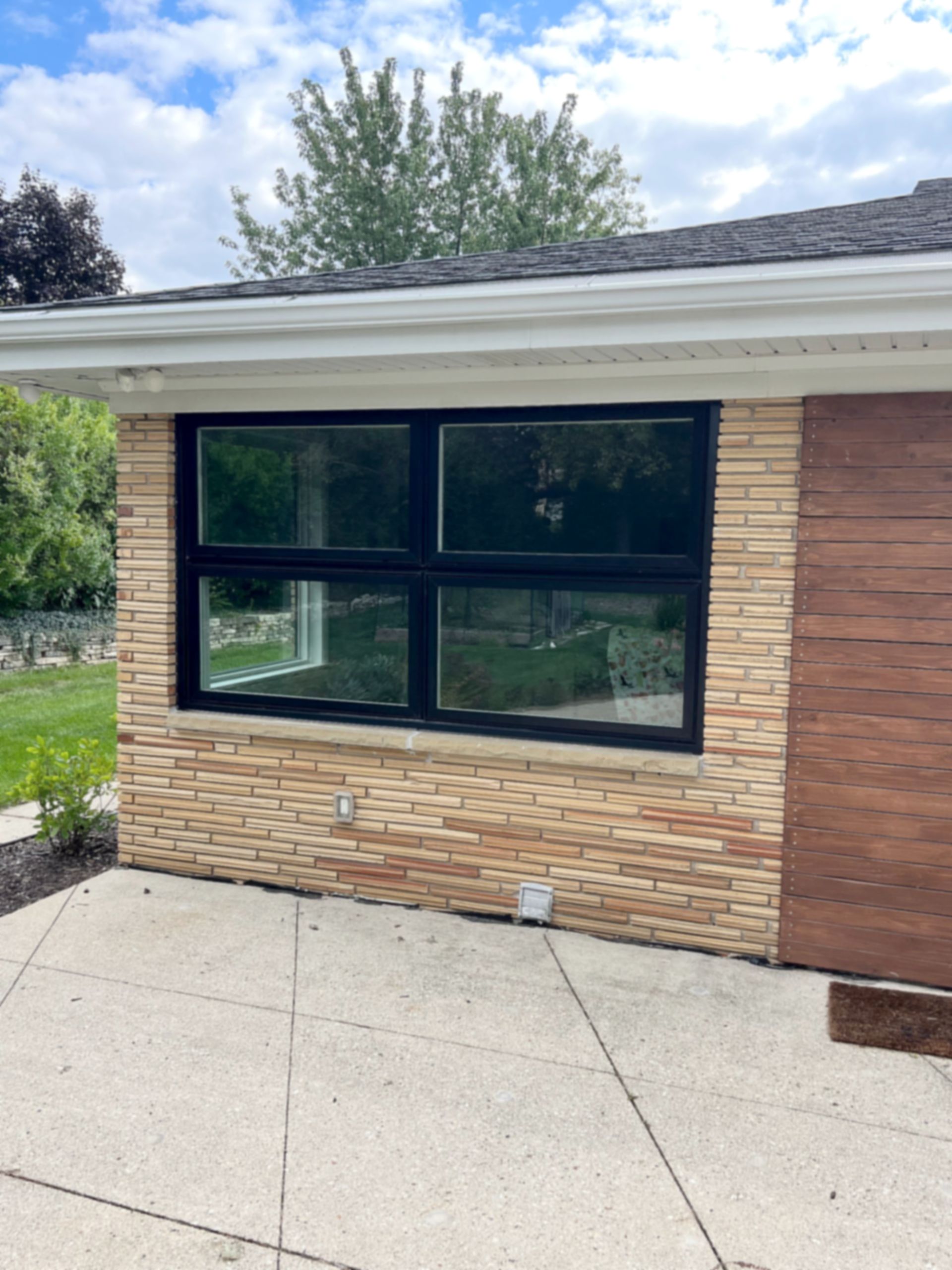 Exterior of a building with a large black-framed window. Beige brick exterior, brown siding, and concrete patio.
