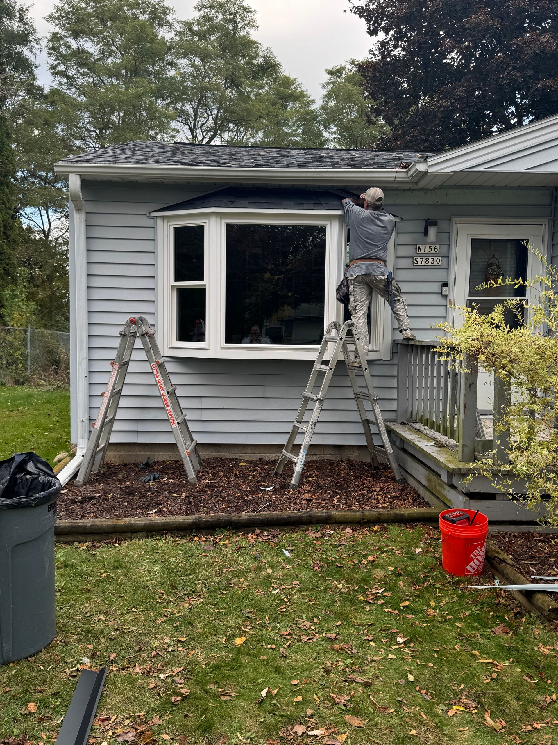 Person on ladder works on a house's exterior; a window and front door are visible, along with tools and a trash can.