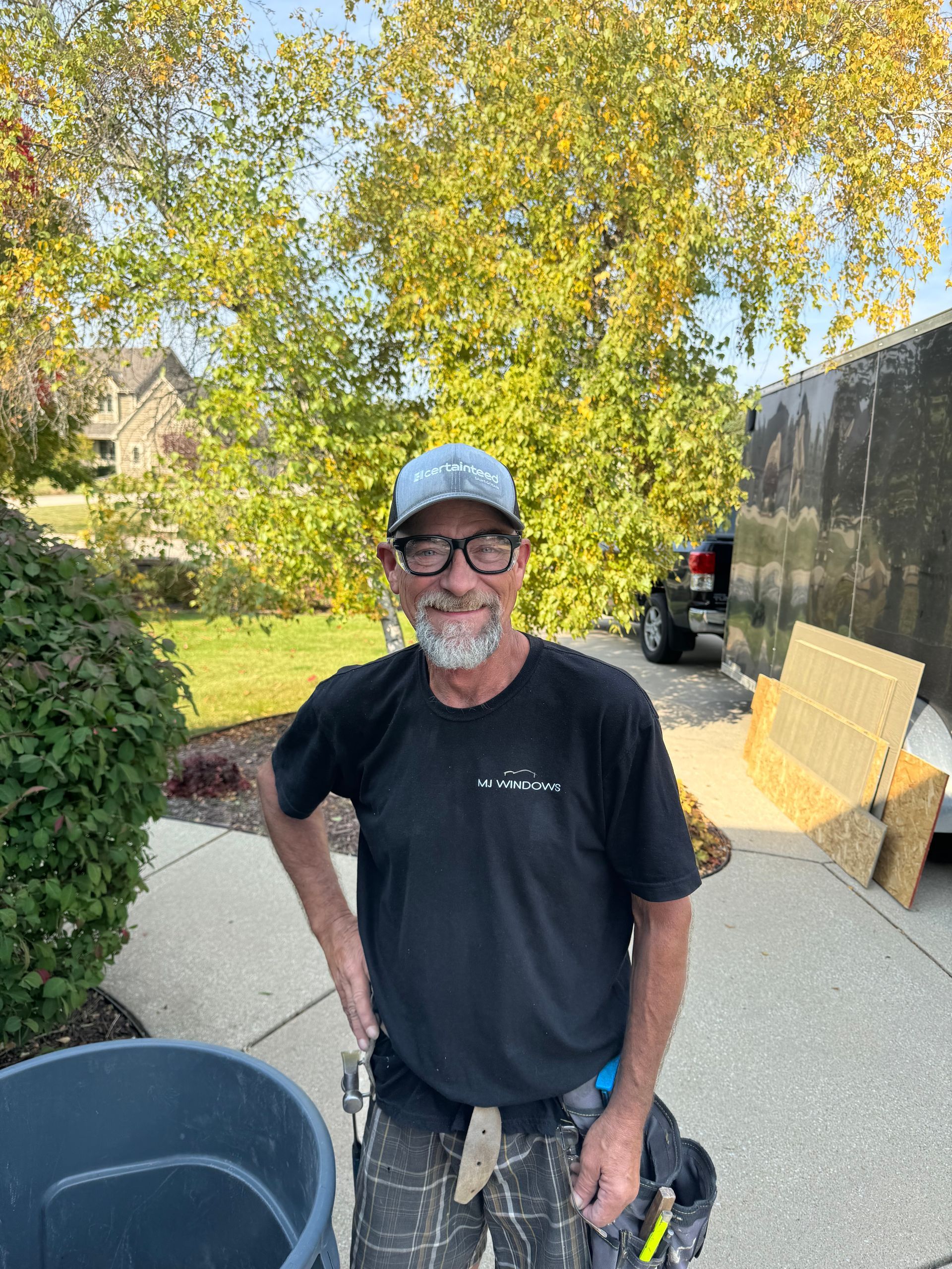 Man in black shirt, cap, standing by a trash can with a trailer and trees in the background.