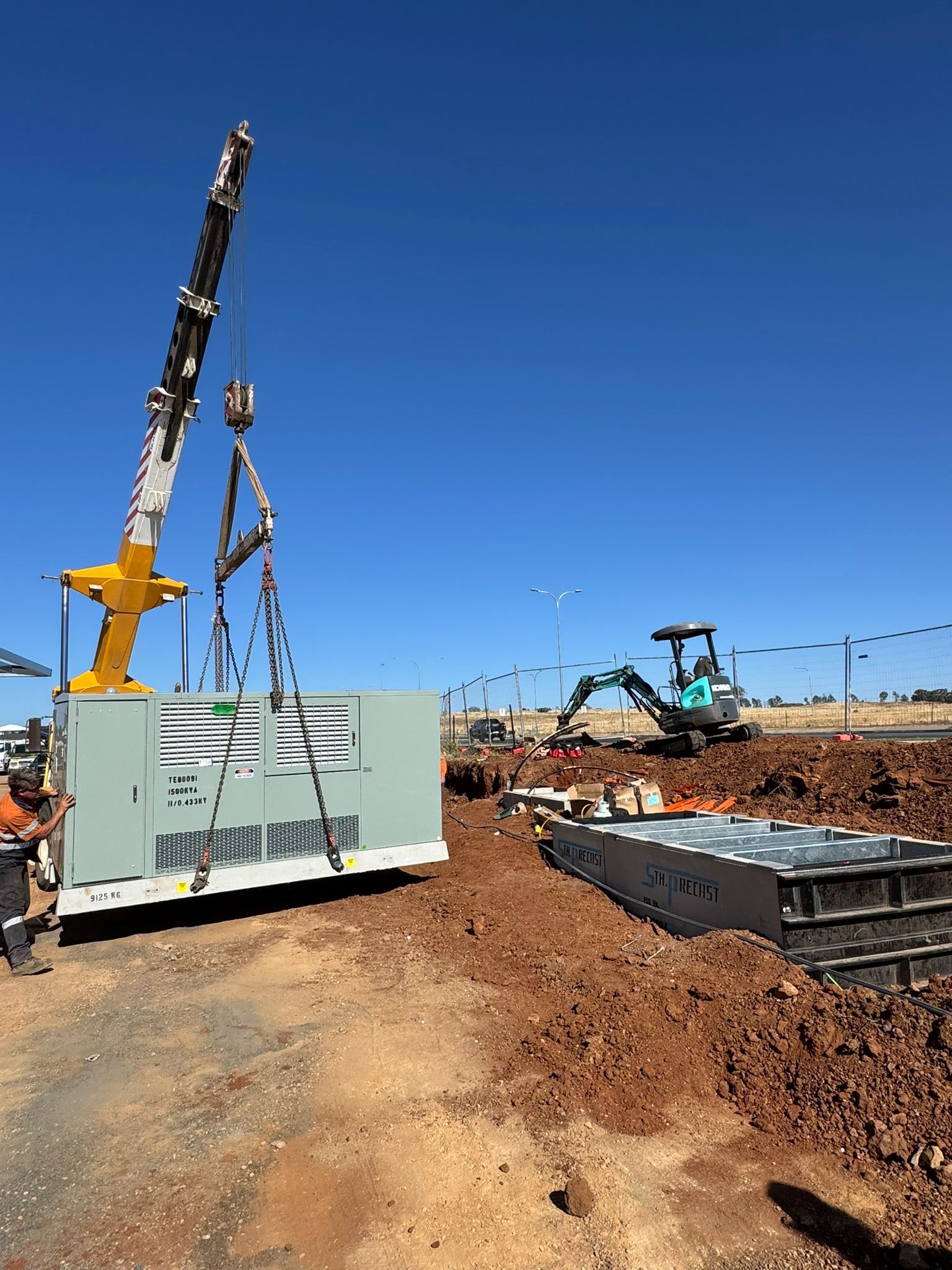 Construction site with crane lifting a white container beside a dirt road under a clear blue sky