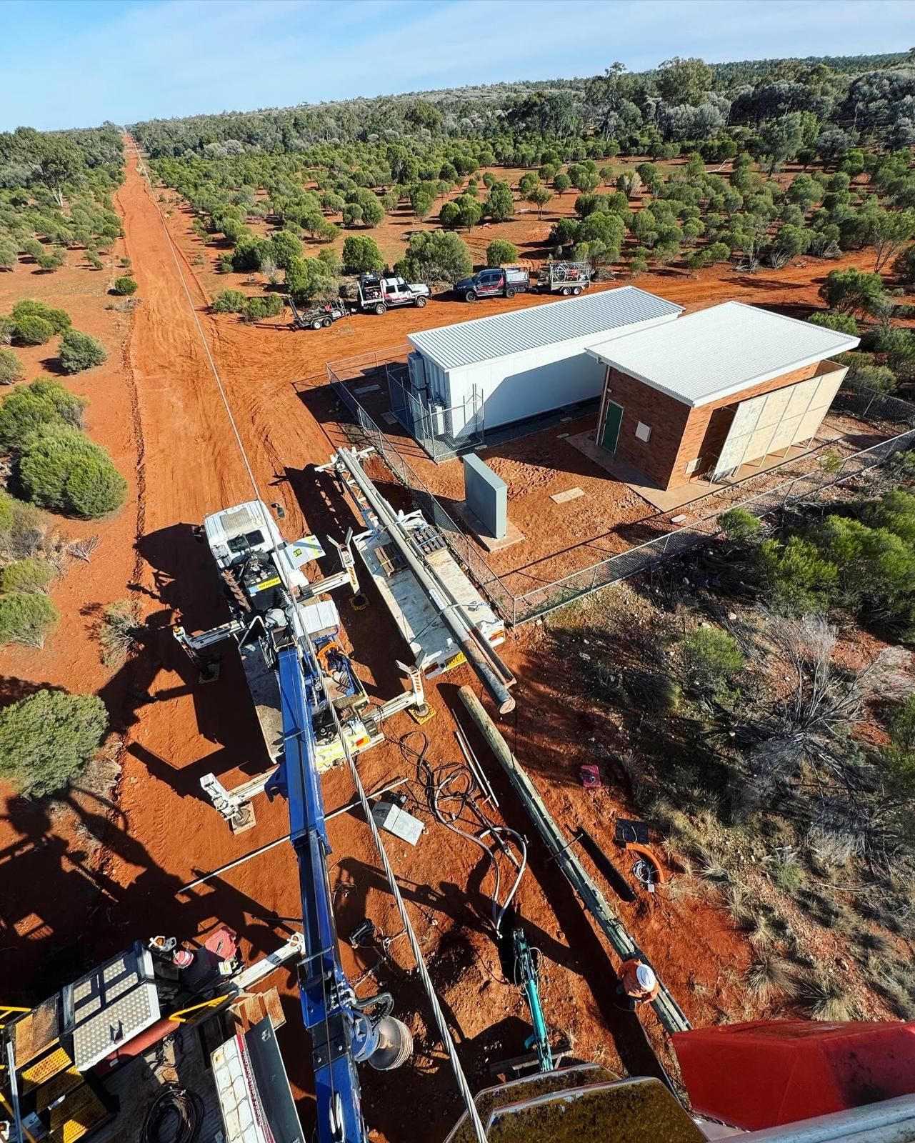 Aerial view of a rural worksite with drilling equipment beside a small building and red dirt road