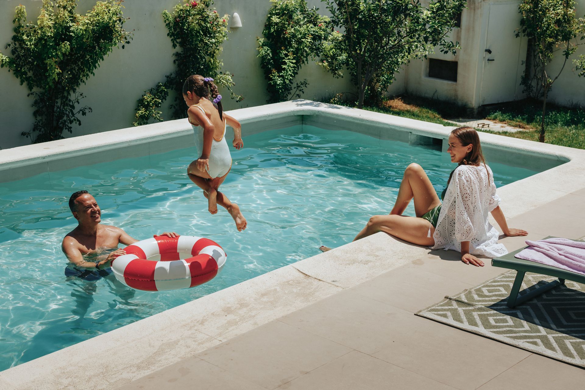 A family of three playing and relaxing by a swimming pool.