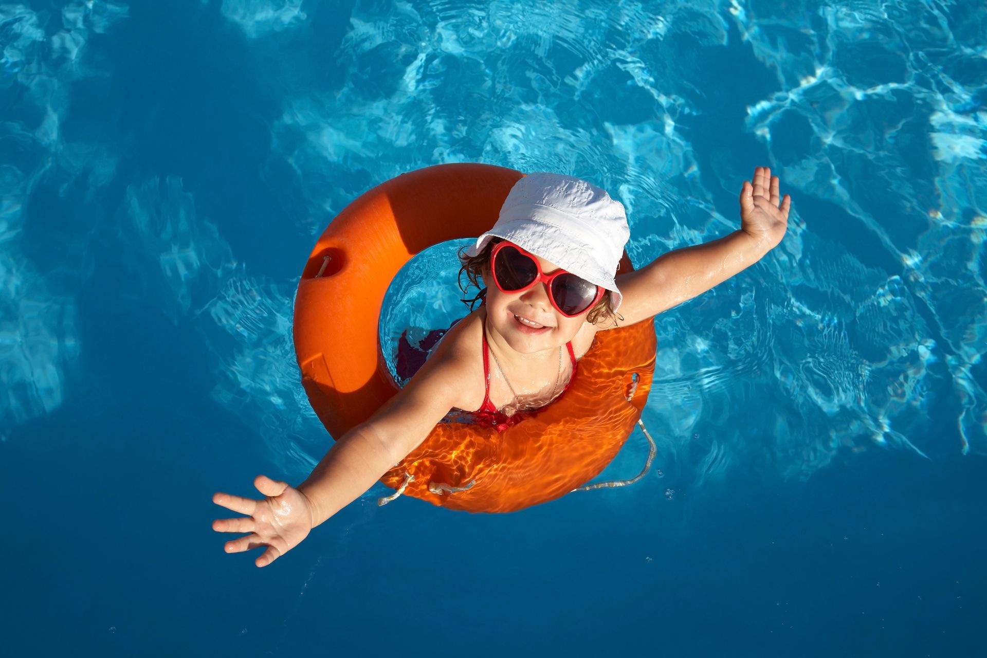 A cheerful little girl enjoys the water in a pool, using an orange life ring for support.