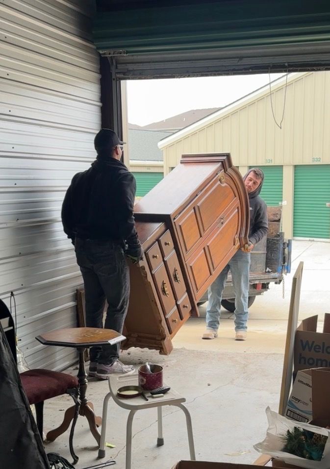 Two workers in matching blue shirts carry a brown sofa toward a truck parked in a suburban driveway near a yellow bin.
