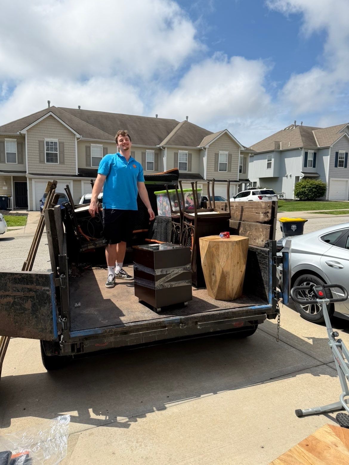 Two people in matching blue shirts carry a brown sofa toward a moving truck on a residential street.