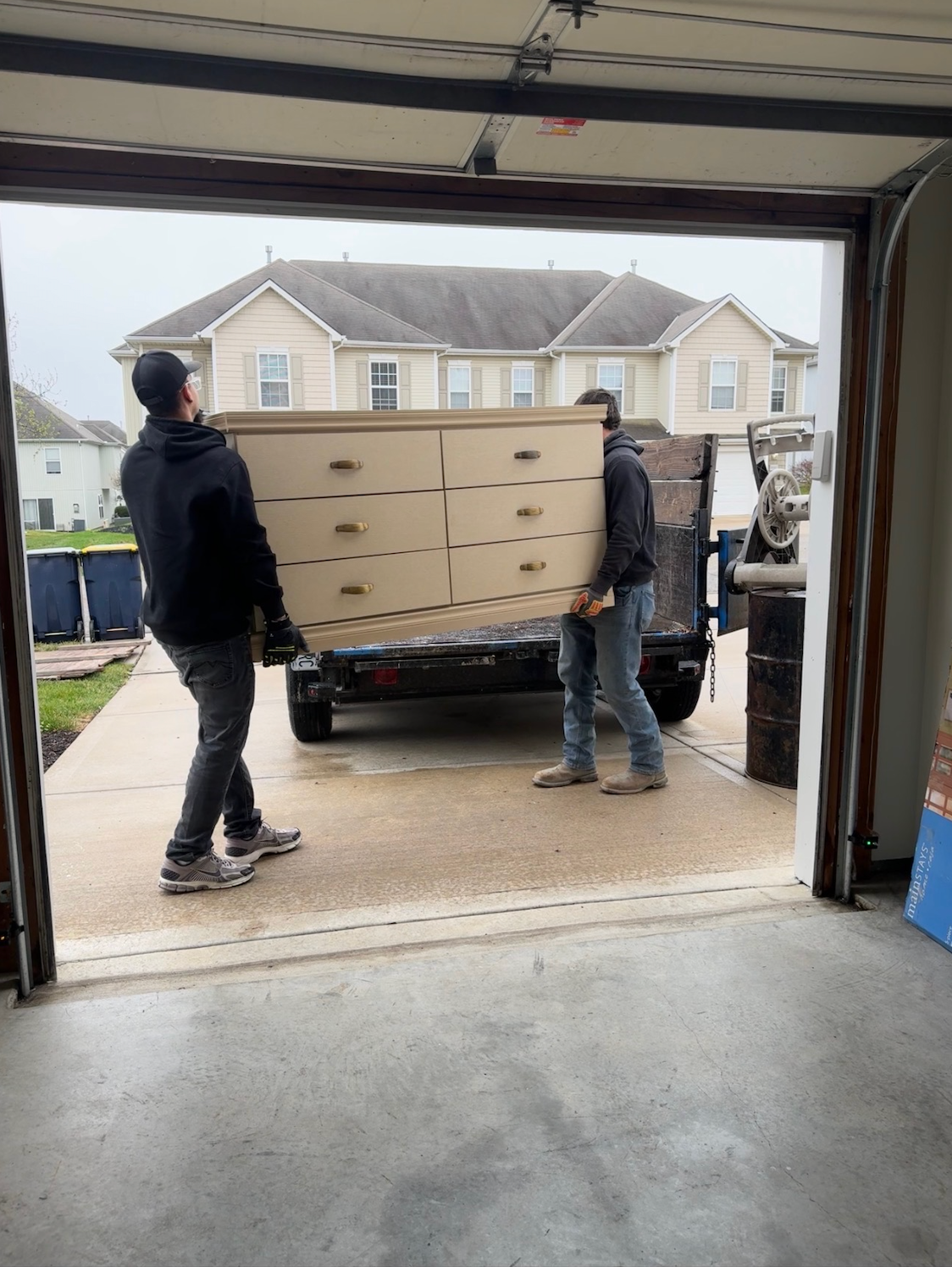 Two people wearing blue shirts with a logo carry old furniture out of a yard filled with clutter and trash bags.