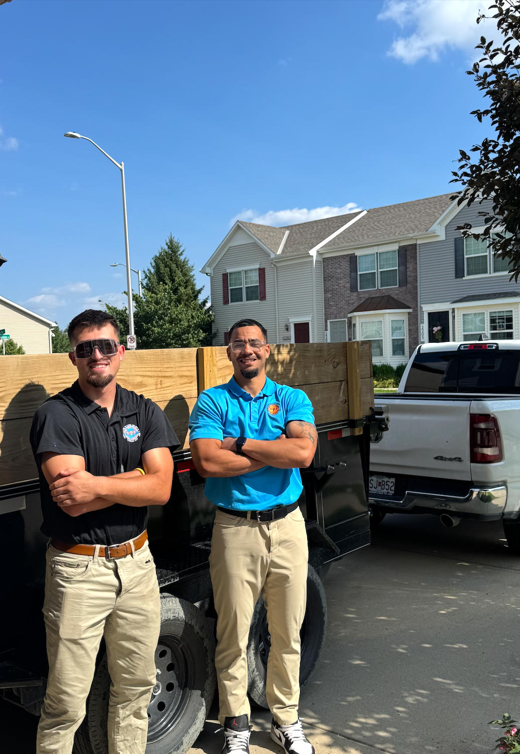 Two movers in blue uniforms carry an armchair toward a moving truck parked in front of a residential house.