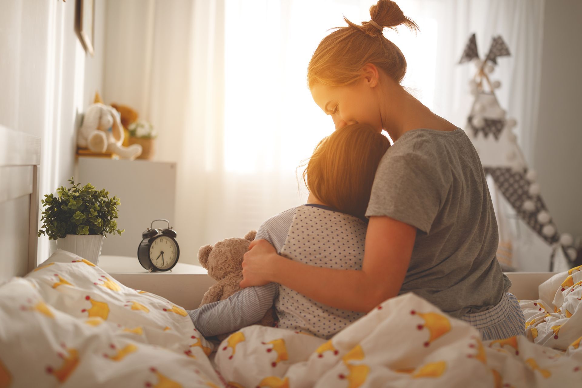 A woman is hugging a child in bed with a teddy bear.