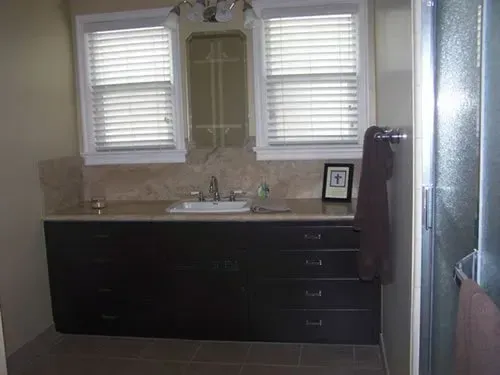 A bathroom with a sink , mirror , drawers and two windows.