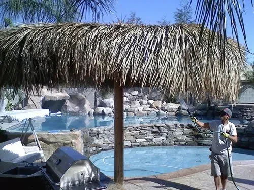 A man is standing under a thatched umbrella next to a pool.