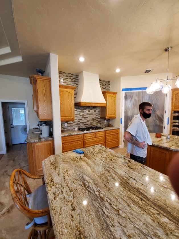 A man wearing a mask is standing in a kitchen with a large granite counter top.