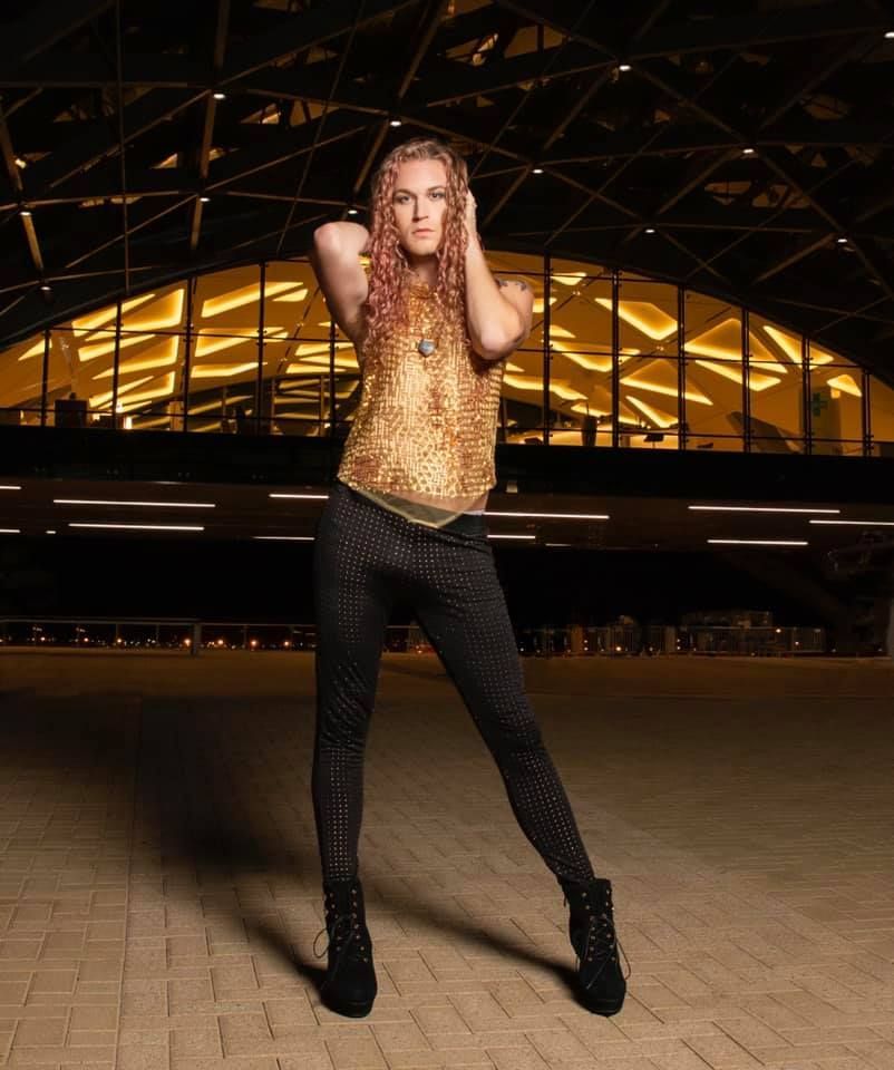 A woman in a gold top and black pants is standing in front of a building.
