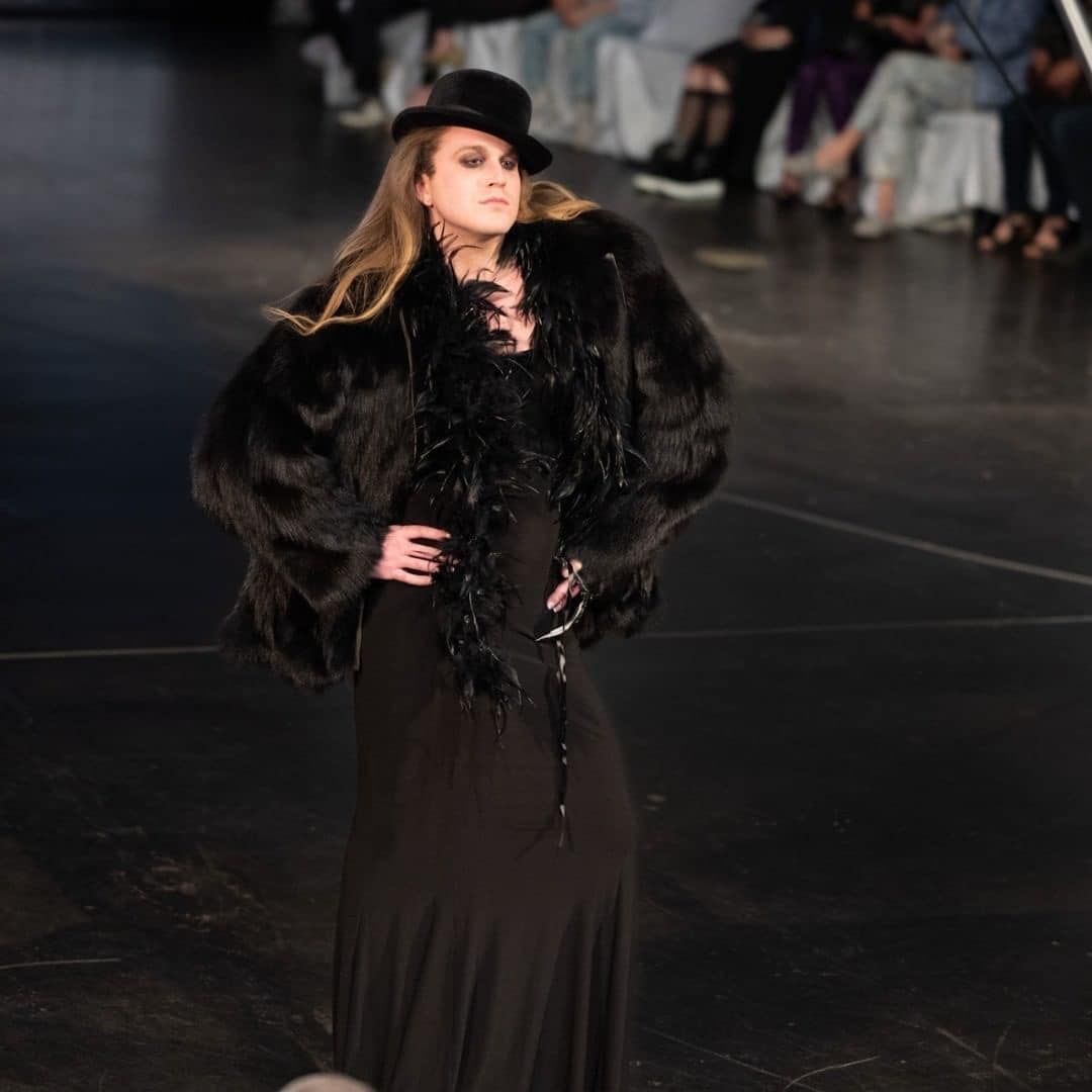 A woman wearing a black fur coat and top hat walking down a runway for a fashion show.