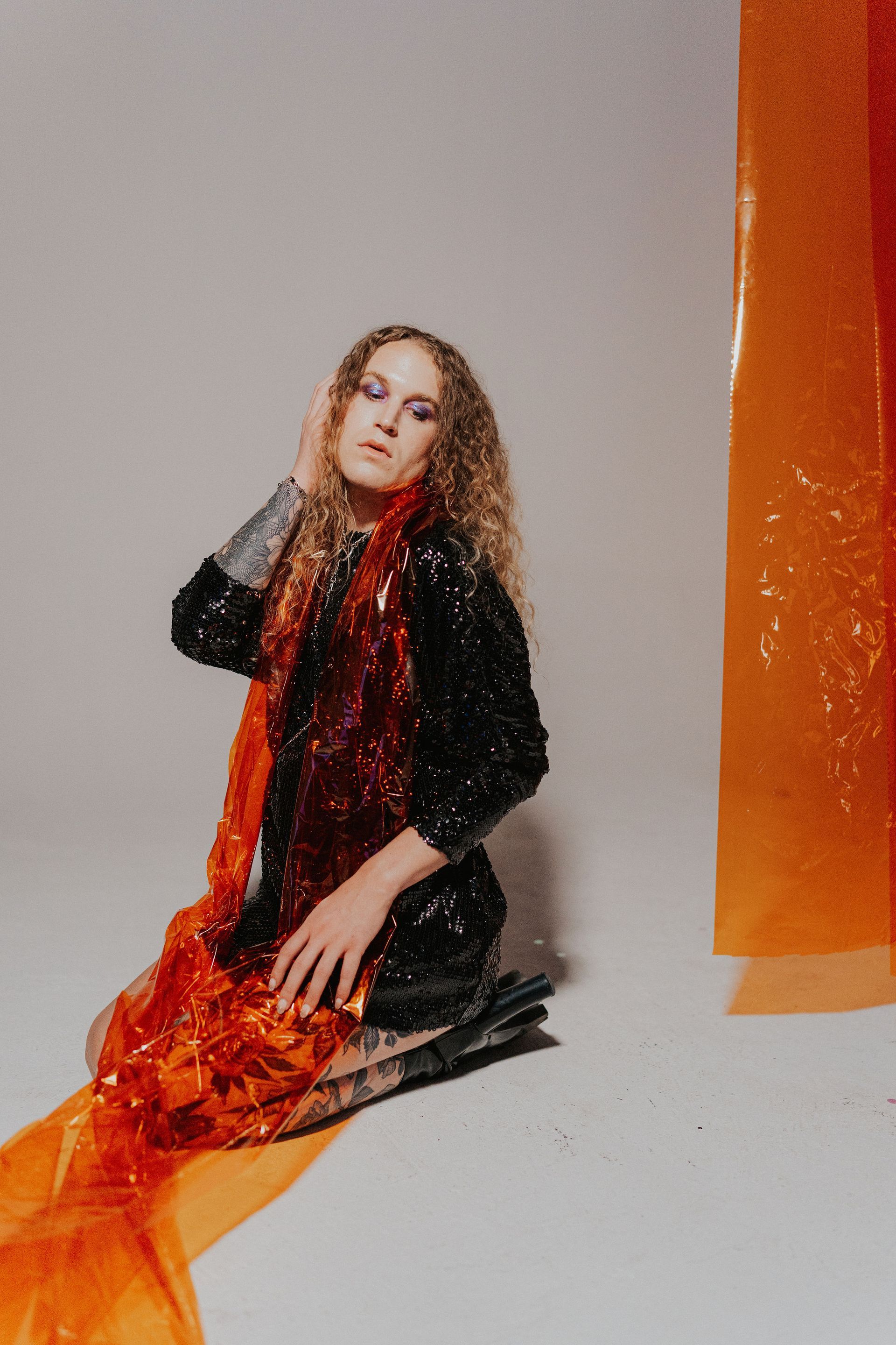 A woman in a black dress is kneeling down in front of an orange curtain for a photo shoot.