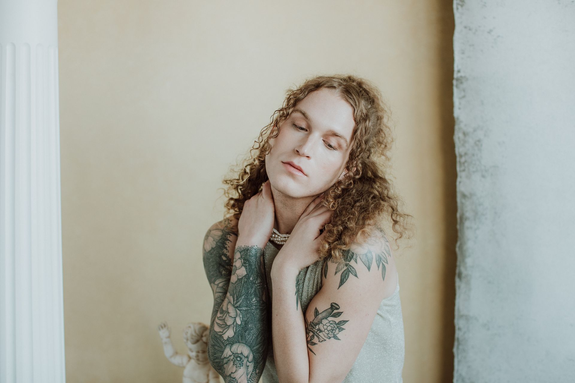 A woman with curly hair and tattoos on her arms is standing in front of a wall for a fashion photo shoot.