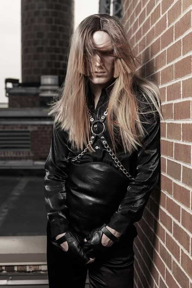 A man with long hair is leaning against a brick wall for a photo shoot.