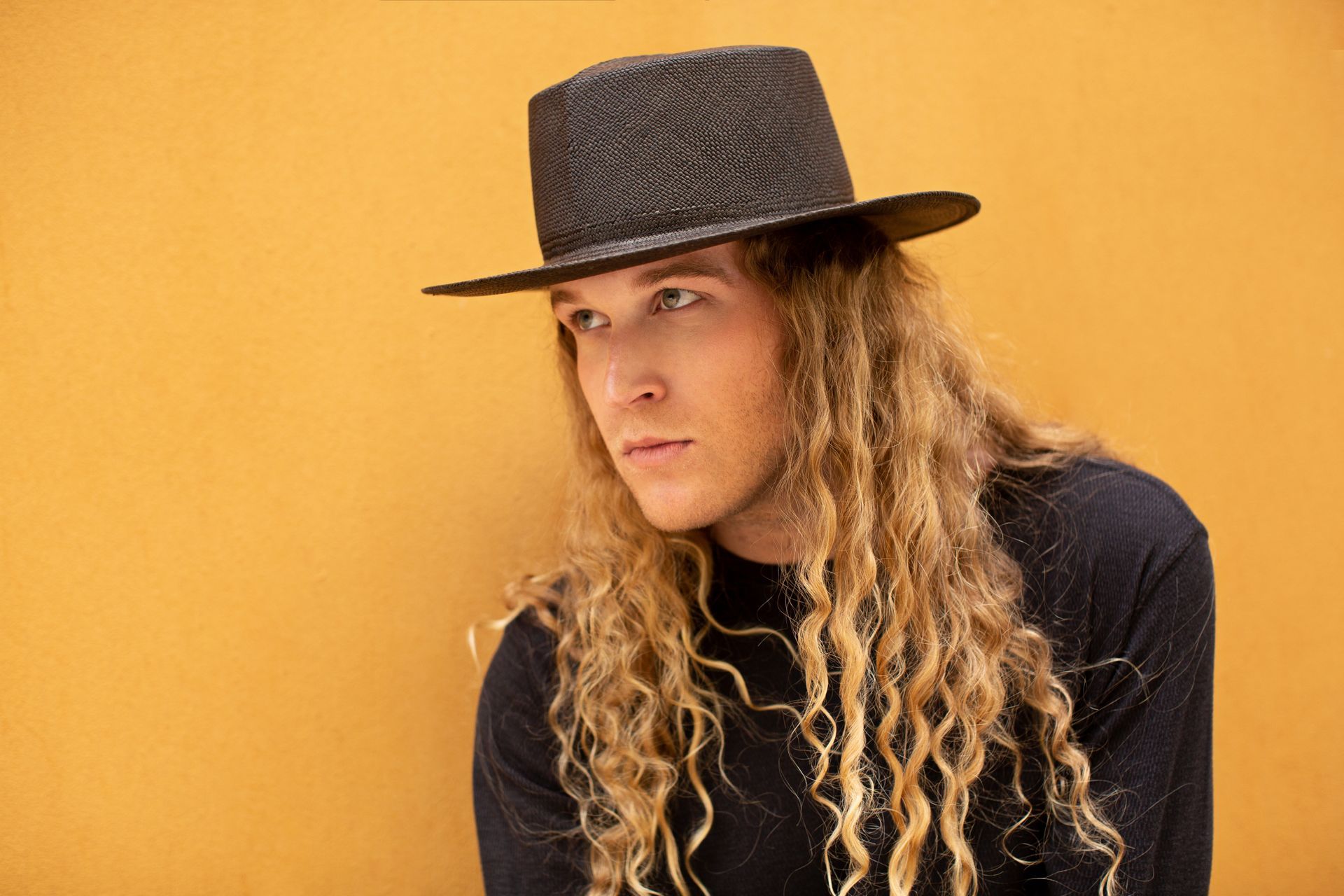 A young man with long curly hair is wearing a black hat for a fashion photo shoot.