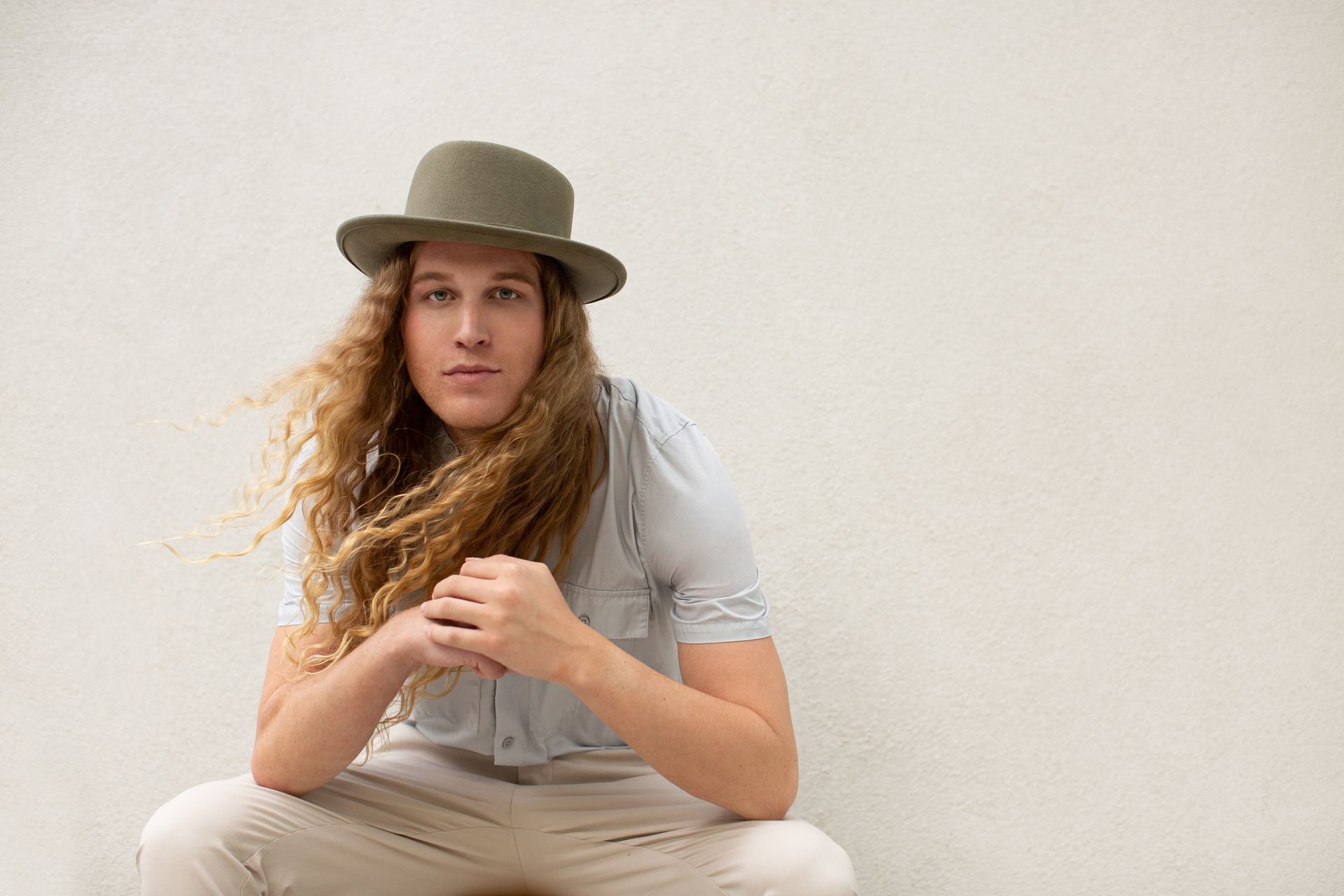 Scotty wearing a hat is kneeling down in front of a white wall for a fashion photo shoot.