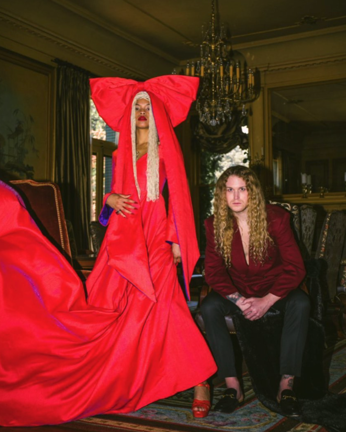 A woman in a red dress is standing next to a man in a chair for a photo shoot.