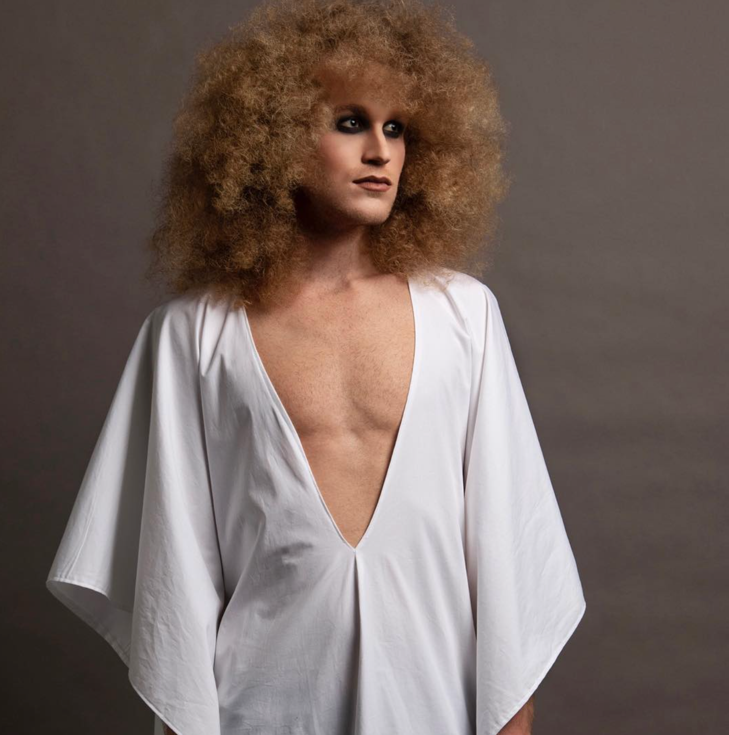 A man with curly hair is wearing a white shirt with a plunging neckline for a photoshoot.