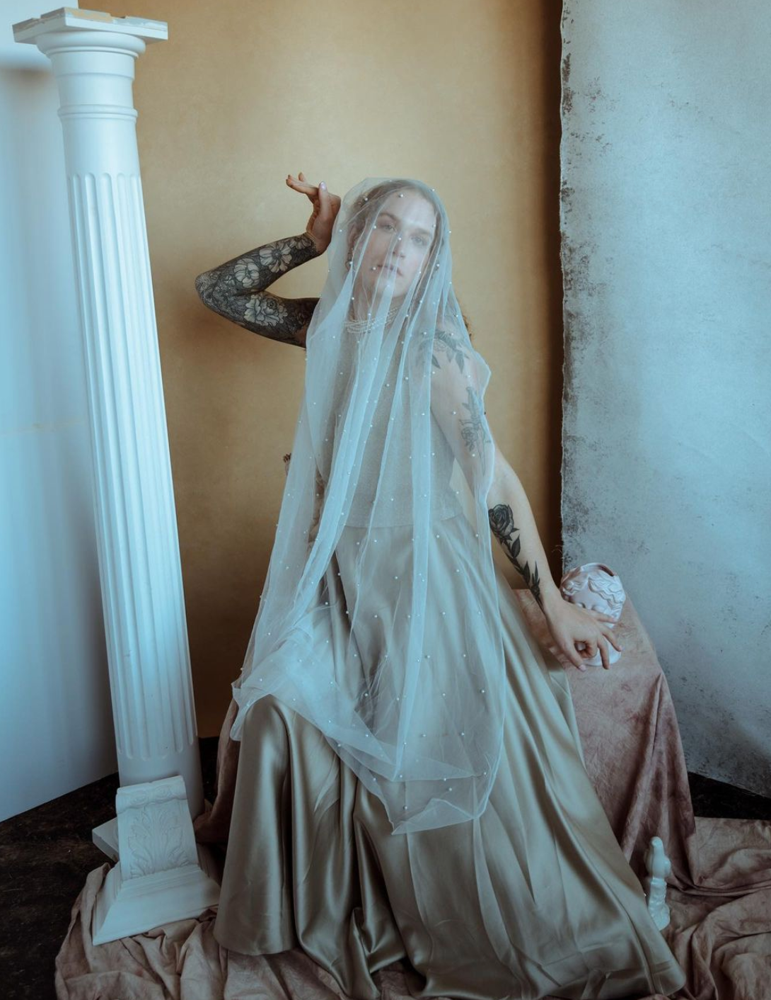 A woman in a long dress is sitting next to a white column for a fashion photo shoot.