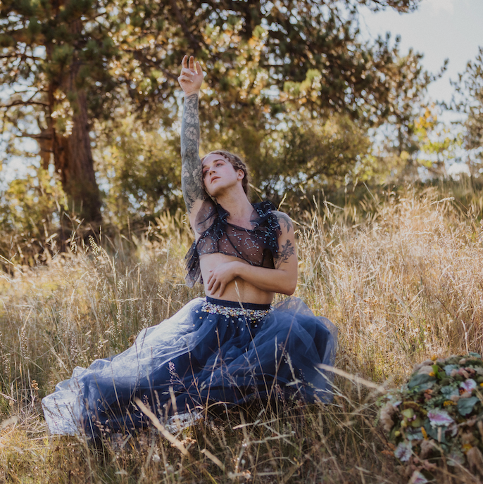 Scotty Strehlow is in a blue dress and sitting in the grass with their arms in the air, posing for the camera.