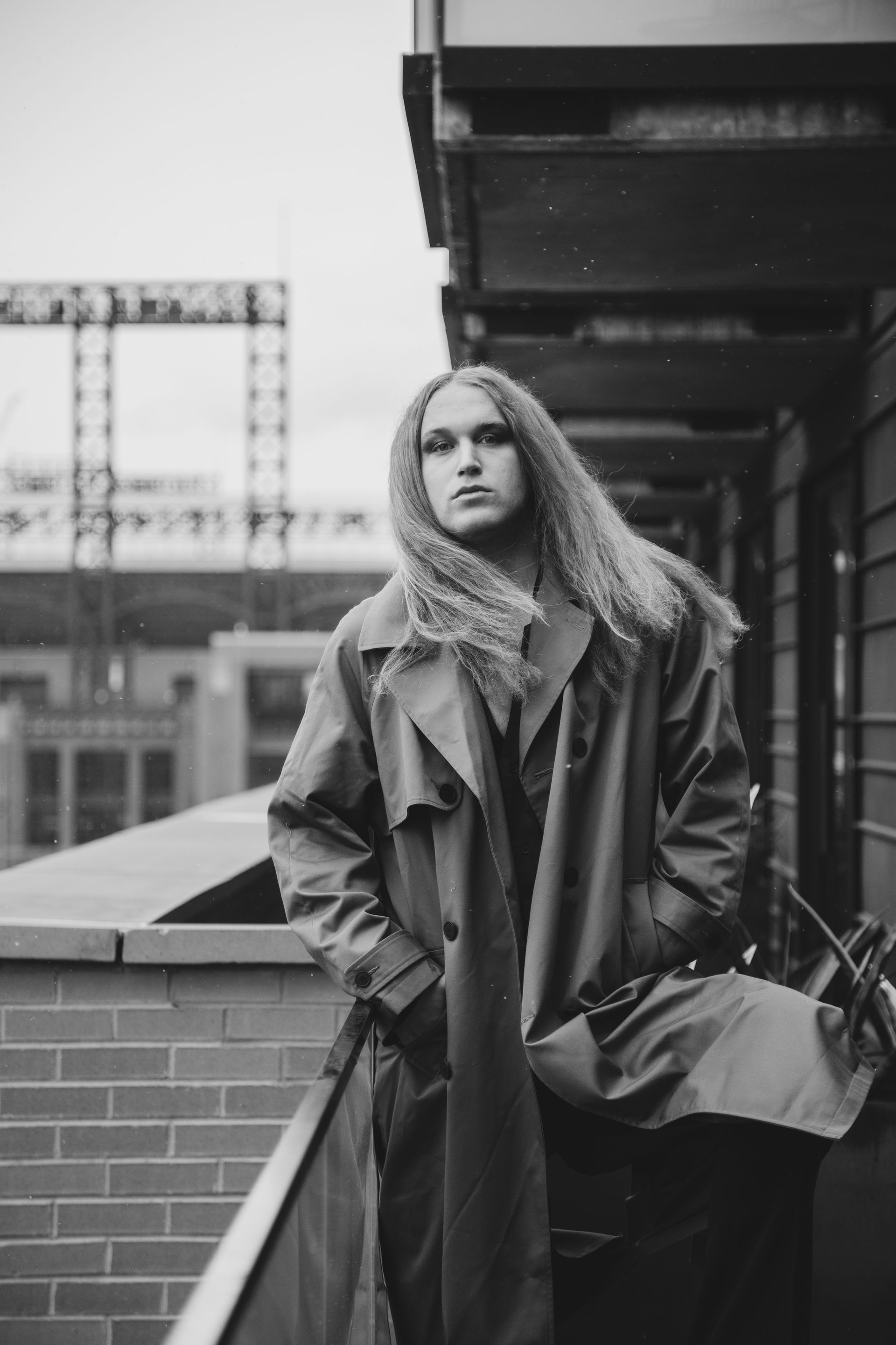 A black and white photo of a man with long hair wearing a trench coat for a fashion photo shoot.
