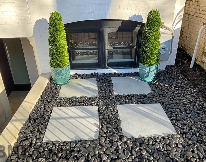 Stone walkway leading to a black fireplace, flanked by tall potted shrubs and dark landscaping rocks