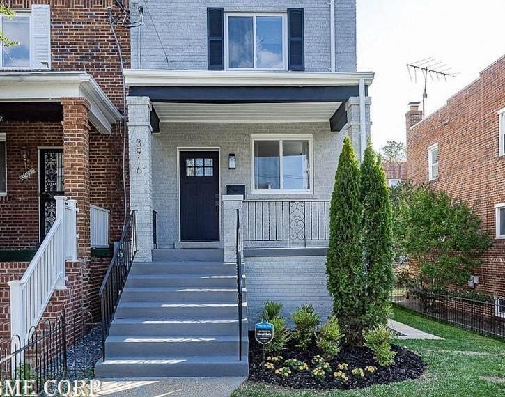 Front view of a brick townhouse with a gray porch, steps, white railing, and two tall evergreens.