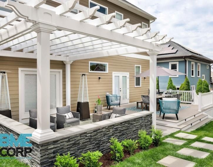 Modern beige house patio with white pergola, gray stone base, outdoor seating, and a small deck by a lawn