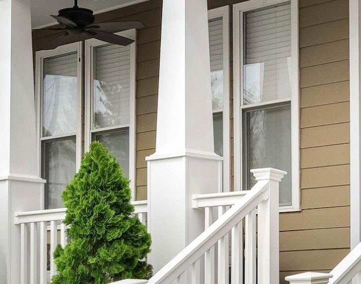 Front porch with white railing, beige siding, and a potted evergreen shrub beneath a ceiling fan