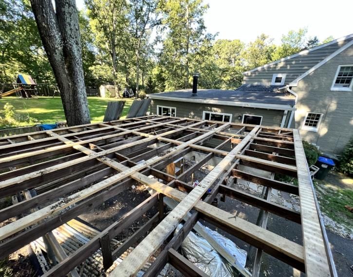 Exposed roof rafters on a house during construction, with trees and a sloped yard nearby.