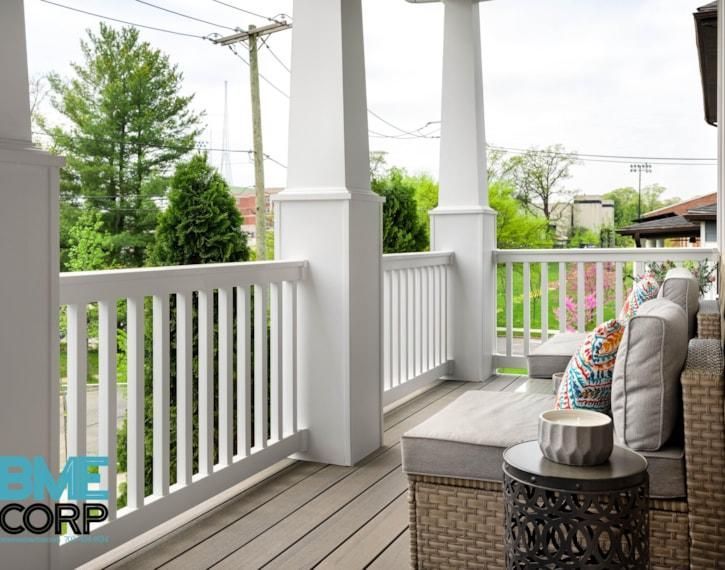 Covered front porch with white railings, gray deck, cushioned bench, and small side table overlooking greenery