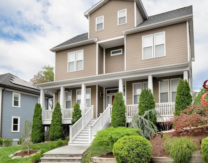 Two-story beige house with white porch railings, front steps, and landscaped shrubs.