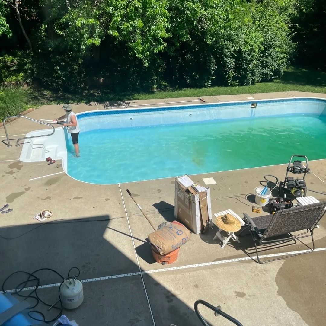 Person cleaning a green-tinted pool. Equipment and supplies are on the concrete deck. Green trees in the background.