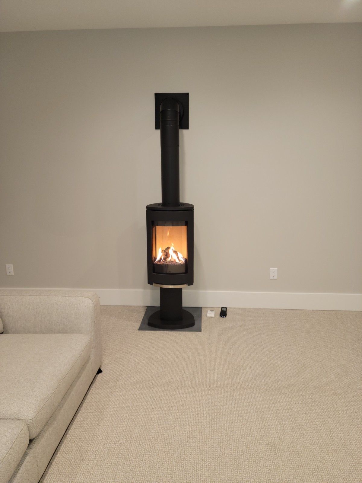 Black wood-burning stove with flames in a neutral-toned room. Fireplace stands on a black base, with a dark flue.