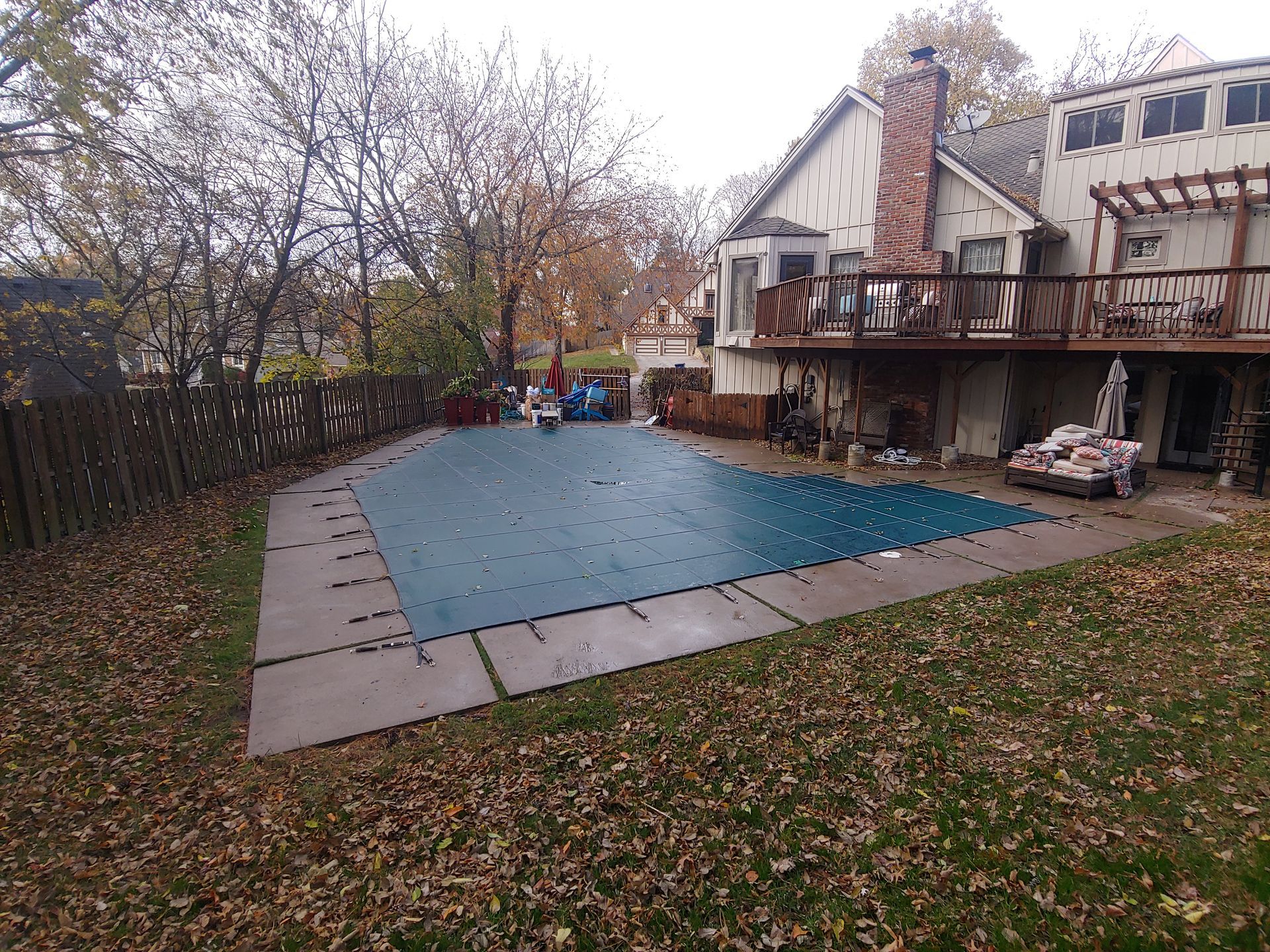 Backyard pool covered with a blue tarp, next to a two-story house with a wooden deck.