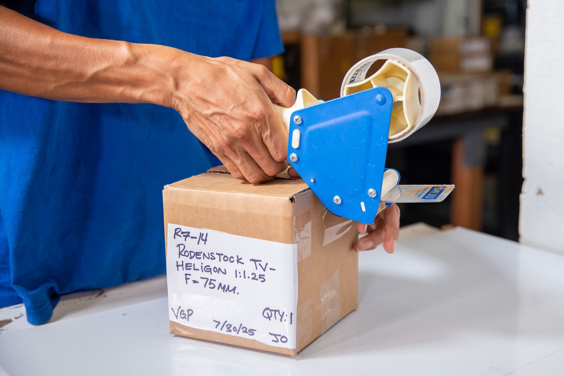 Hands sealing a cardboard box with blue packing tape dispenser on a white table