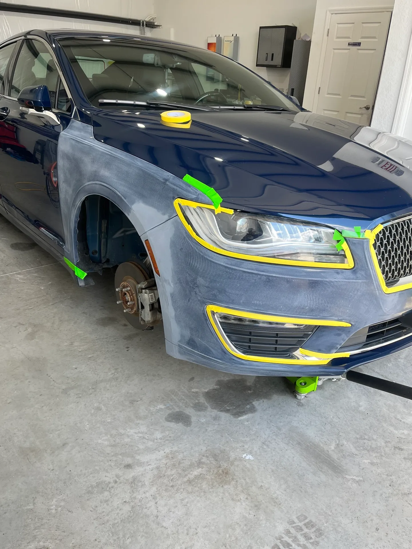 Blue car in a repair shop with front-end damage. Bodywork partially sanded, fender removed, on green stands.