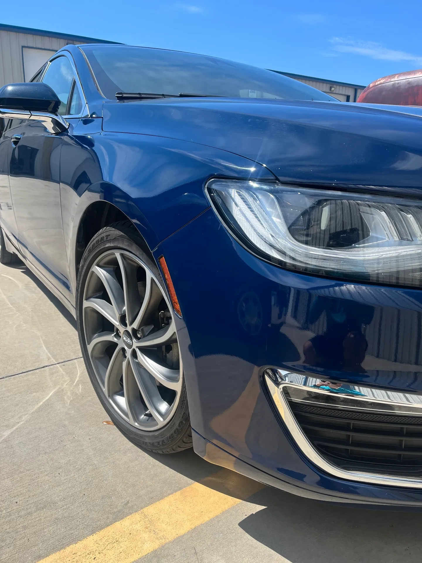 Blue Lincoln MKZ car parked outdoors, close-up of front. Shiny paint, silver rims, sunny day.